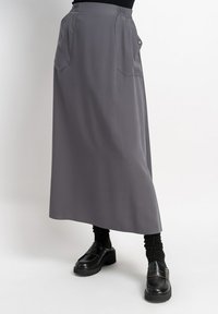 Langer grauer Rock aus glatt gewebtem Stoff, mit zwei frontalen Taschen und einem elastischen Bund für zusätzlichen Komfort. Getragen mit schwarzen Plateauschuhen.