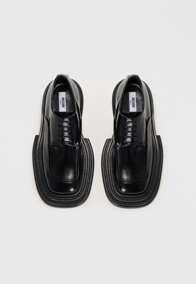 Pár lesklých černých kožených šněrovacích bot s vysoce zesílenými podrážkami a čtveratými špičkami, pohled shora na bílém pozadí.