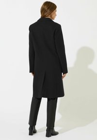 Cappotto di lana nera con design classico, dotato di uno spacco posteriore, maniche lunghe e colletto a punta. Abbinato a pantaloni neri sartoriali e stivaletti alla caviglia.