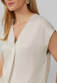 Beige, mouwloos topje van zachte stof met een V-hals en subtiele draping, voorzien van een gladde textuur en zijsplitten voor comfort.