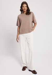 Luźna, taupe bluzka z krótkim rękawem i okrągłym dekoltem, zestawiona z białymi spodniami o prostym kroju i brązowymi mokasynami. Gładka tekstura, casualowy wygląd.