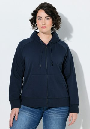 Kvinde med kort krøllet hår iført øreringe med ringe, en mørkeblå hættetrøje med lynlås og blå jeans mod en ensfarvet lys baggrund.