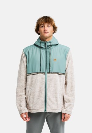 Giovane uomo che indossa una giacca leggera beige chiaro e turchese con zip, cappuccio e rifinitura decorata, in piedi davanti a uno sfondo semplice.