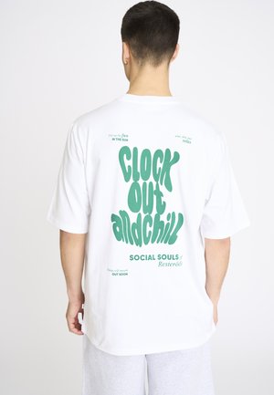Mand iført hvid oversized T-shirt med grøn tekst "Clock out and chill" og afslappede lysegrå shorts, stående mod en hvid baggrund.