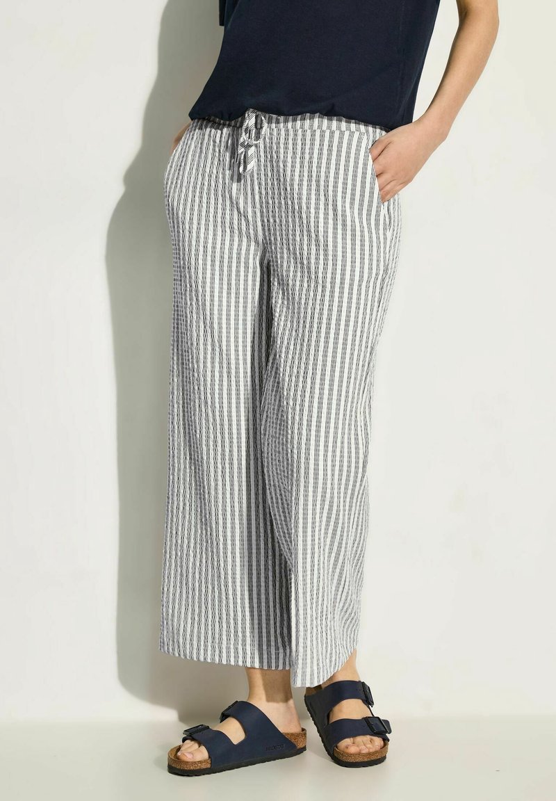 Striped coastal pants in white and navy, loose fit, mid-rise with drawstring waist, featuring two side pockets. Worn with dark sandals.