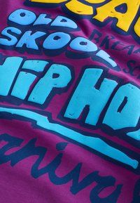 Gros plan d'un tissu violet avec un texte en relief coloré incluant "old skool hip hop" en lettres stylisées bleues et jaunes.