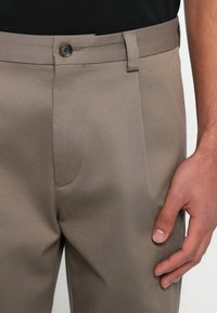 Μπεζ παντελόνι σε ίσια γραμμή με λεία υφή, διαθέτει κούμπωμα με κουμπί, διακριτικές πιέτες και θηλιές για ζώνη. Χέρι ακουμπά στην περιοχή της μέσης.