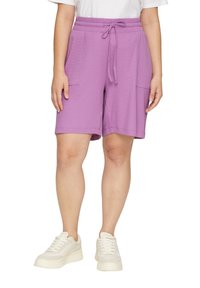 Lila Shorts aus strukturiertem Stoff, mit einem elastischen Bund und einem Kordelzug sowie zwei Vordertaschen. Getragen mit weißen Turnschuhen.