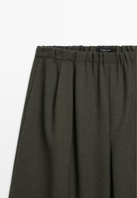 Pantaloni verdi scuri a vita alta realizzati in tessuto texturizzato con pieghe davanti e una cintura elastica. Etichetta visibile all'interno.