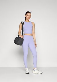Světle fialový sportovní crop top a odpovídající legíny s vysokým pasem. Černá kabelka na rameno a bílé tenisky doplňují tento outfit. Hladká textura.