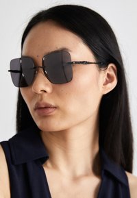 Ochelari de soare negri, supradimensionați, cu lentile pătrate, cu rame subțiri negre și accente cu modele complexe pe temple. Purtați de un model cu părul lung și negru.