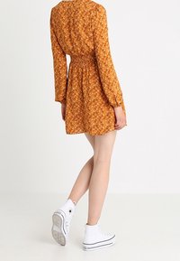 Oranssi kukkamekko, jossa on pitkät hihat ja smockattu vyötärö, yhdistettynä valkoisiin korkeisiin lenkkitossuihin. Materiaalina kevyt, teksturoitu kangas.