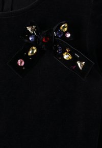 Black velvet fabric with a decorative bow featuring colorful gemstones: red, yellow, blue, and pink. Textured with a shiny finish.