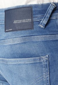Pantalones de mezclilla de color azul claro con una etiqueta de cuero que dice "DENIM TOM TAILOR" y "DENIM DESIGN STUDIO". Se aprecian detalles de costura notables y bolsillos visibles.