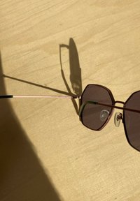 Lunettes de soleil hexagonales avec verres teintés marron, une fine monture en métal doré rose, et des embouts de branches accentués de vert. Ombre légère projetée sur une surface beige.