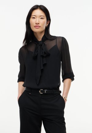 Femme aux longs cheveux noirs portant un chemisier noir transparent avec un nœud noué au col et un pantalon noir, les mains dans les poches.