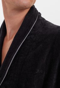 Accappatoio in peluche nero con tessuto texturizzato, dotato di colletto a scialle e piping grigio. Branding discreto visibile sulla parte sinistra anteriore.