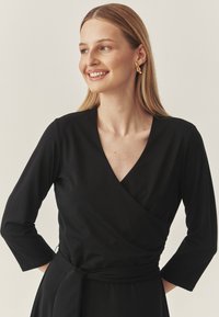 Zwarte wikkeljurk met lange mouwen, V-hals en een ceintuur in de taille. Gladde stoftextuur, minimalistisch ontwerp, die ingetogen elegantie benadrukt.