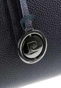 Primo piano di una borsa in pelle nera testurizzata con un charm rotondo argento lucido del logo Pierre Cardin attaccato con una tracolla in pelle.
