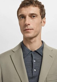 Beige blazer over a dark gray button-up polo. Blazer features a notched lapel and a pocket square. Smooth fabric, tailored fit.