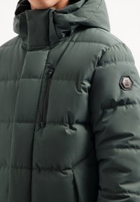 Donkergroen gewatteerde jas met een hoge kraag, zwarte ritszak, horizontale stiknaden en een hexagonaal logopatch op de schouder.