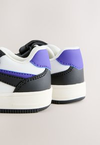 White sneakers with black and purple accents, thick white soles, and padded collars against a light neutral background.