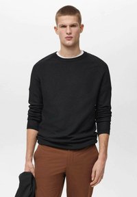 Črn teksturiran pulover z dolgimi rokavi, okroglo ovratnico in elastičnimi manšetami, v kombinaciji z rjavimi hlačami. Ozadje je enostavno belo.