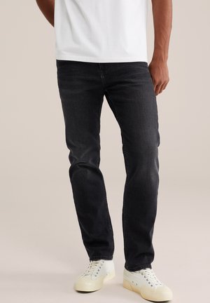 Homme portant un jean noir slim, des baskets blanches et un t-shirt blanc, debout devant un fond neutre.