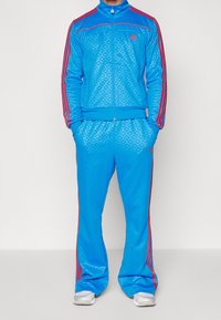 Modrý teplákový komplet s texturovaným vzorem, červené proužky na rukávech a kalhotách, bunda na zip a odpovídající volné kalhoty.