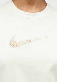 Κοντινή λήψη λευκού T-shirt με κεντημένο λογότυπο Nike σε μπεζ χρώμα στη μέση του στήθους.