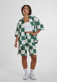 Camicia a maniche corte a scacchi verdi e bianchi abbinata a pantaloni corti coordinati. Entrambi hanno una vestibilità comoda e una texture morbida; indossati con sneakers bianche.