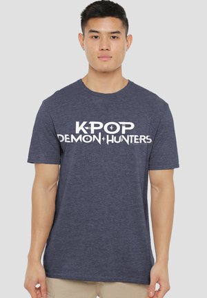Junger Mann, der ein dunkelblaues T-Shirt mit weißem Aufdruck "K-POP DEMON HUNTERS" trägt, steht vor einem einfachen hellen Hintergrund.