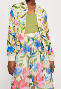 Blomstret bomberjakke i letvægtsstof med multikolorede blomsterprint, med lynlås foran og ribbede manchetter, parret med en grøn top.