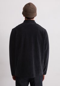 Zwarte corduroy jas met lange mouwen, met een ontspannen pasvorm en een kraag aan de achterkant. Draagt een bruine gebreide beanie.