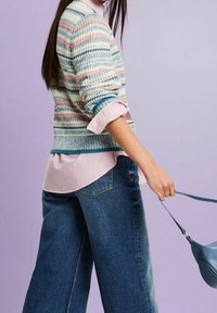 Femme portant un pull rayé sur une chemise rose et un jean large, tenant un sac à main bleu devant un fond lavande.