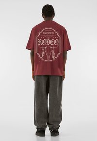 Maroon oversized t-shirt featuring a graphic design with "RODEO" and skull accents. Worn with grey wide-leg pants and black sneakers.