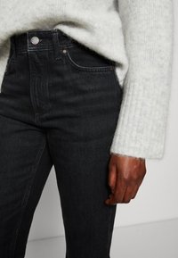 Lähikuva henkilöstä, joka käyttää mustia farkkuja ja vaaleanharmaata pörröistä neuletta, näyttäen vyötärön ja oikean käden rentona sivulla.