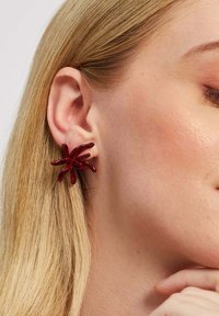 Červené akrylové náušnice v tvare voľného pavúka, s predĺženými, zakrivenými nohami, lesklým povrchom a bezpečným uchytením na laloku ucha.