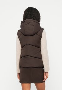 Gilet imbottito marrone con cappuccio, caratterizzato da un design a zigzag, indossato sopra una maglietta a maniche lunghe di colore chiaro e una gonna scura.