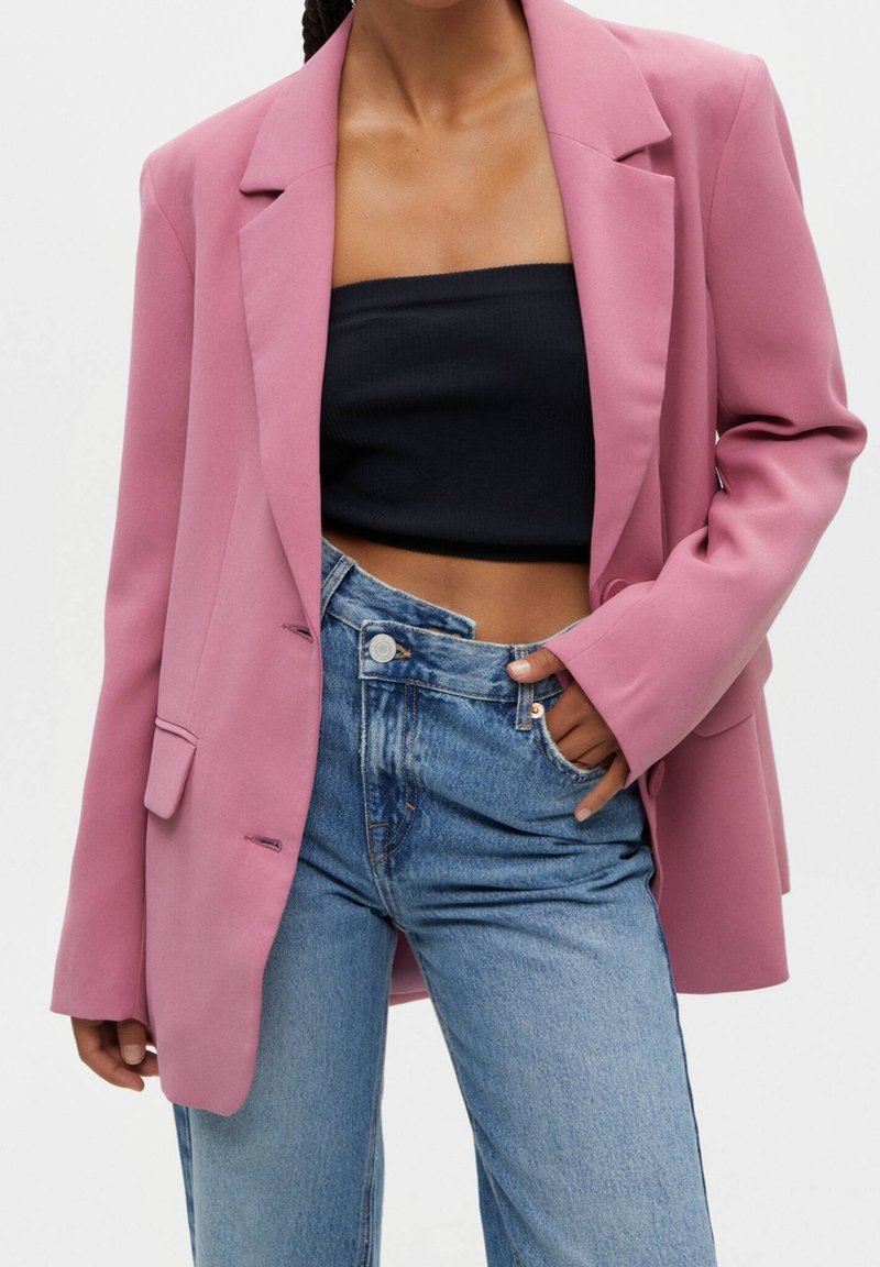 Blazer rose sur mesure en tissu lisse, doté de revers crantés, d'une fermeture à un bouton et de poches plaquées, associé à un jean bleu taille haute.