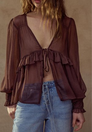 Nuori nainen pukeutuneena läpikuultavaan ruskeaan puseroon, jossa on röyhelöitä ja edessä solmittava nauha, yhdistettynä korkeavyötäröisiin sinisiin farkkuihin, joissa on nyöritys vetoketjun sijaan.