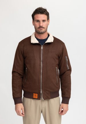 Man met een donkerbruine bomberjas met schapenvachtkraag, beige broek en een donkerblauw overhemd, staand tegen een effen witte achtergrond.