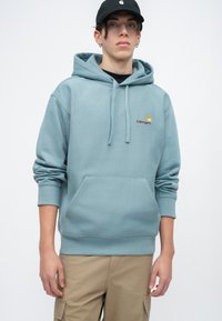 Hanorac din bumbac bleu deschis, cu buzunar frontal, glugă cu șnur și logo Carhartt în galben și bordeaux. Asortat cu pantaloni cargo bej.