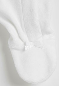 Chaussons en tissu blanc et moelleux avec un bout rond et un petit nœud en satin blanc à la cheville. Texture douce, design simple.