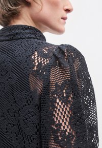Top in pizzo nero con motivi intricati, maniche testurizzate e collo alto, che presenta una combinazione di elementi solidi e trasparenti.
