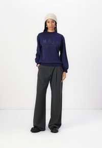 Sweatshirt azul marinho com estampa de horizonte urbano, combinado com calças largas cinza-escuras e sapatos pretos robustos; usado com um gorro de malha bege.