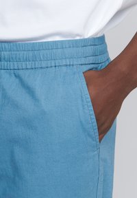 Pantaloni albaștri deschiși cu talie elastică, având un buzunar lateral și o textură netedă, moale. Materialul pare ușor și respirabil.