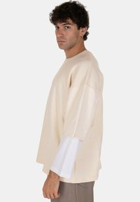 T-shirt beige oversize con un fit rilassato, dotata di maniche corte e un dettagliato orlo bianco a strati. Texture morbida con un design minimale.