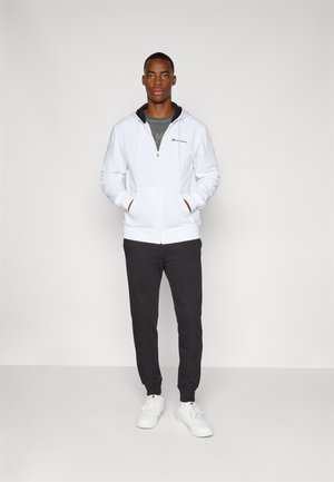White zip-up hoodie with a drawstring, black accents, gray T-shirt underneath, paired with dark sweatpants and white sneakers.