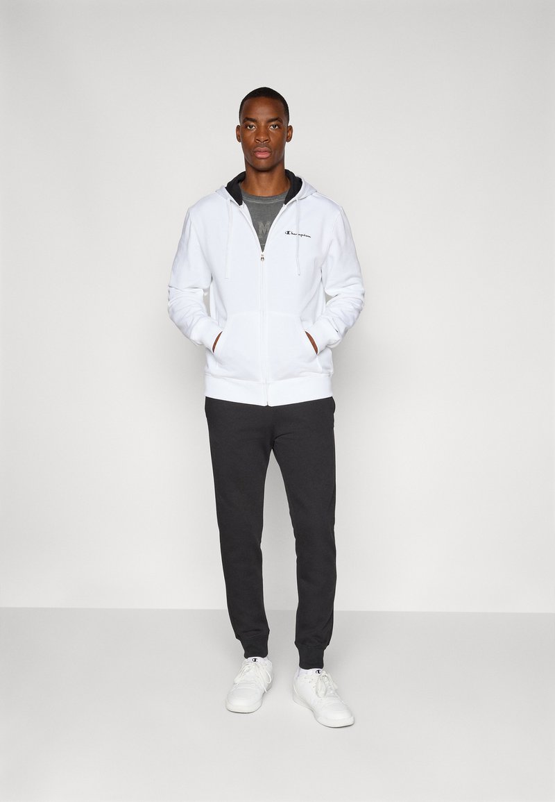 Witte rits-hybride hoodie met een trekkoord, zwarte accenten, grijs T-shirt eronder, gecombineerd met donkere trainingsbroeken en witte sneakers.